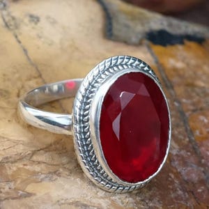 May include: A silver ring featuring an oval, faceted, deep red gemstone. The gemstone is set in a decorative bezel with a braided design. The ring is displayed on a textured, brown surface.