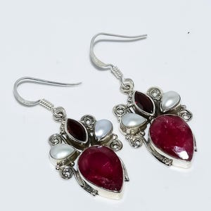 Könnte beinhalten: Silberne Ohrringe mit einem tropfenförmigen, tiefroten Edelstein, ergänzt durch weiße Perlen und dunkelrote Steine. Die Ohrringe haben ein dekoratives, wirbelndes Design.