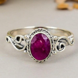 Op de afbeelding: Een zilveren ring met een ovale, dieproze edelsteen. De edelsteen is gezet in een touwachtige bezel. De band van de ring heeft decoratieve krullen aan weerszijden van de edelsteen. De ring is een sieraad.