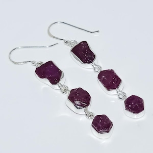 May include: A pair of long drop earrings featuring multiple raw, deep magenta-coloured stones set in polished silver-toned metal. Each earring consists of three vertically linked, irregularly shaped stones suspended from a simple silver hook wire.
