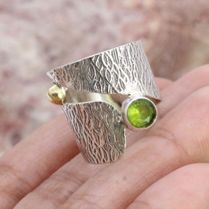 Könnte beinhalten: Ein breiter Silberring mit einer strukturierten, rindenartigen Oberfläche. Der Ring ist offen gestaltet, wobei eine Seite mit einer kleinen, glatten goldfarbenen Kugel und die andere Seite mit einem runden, facettierten, hellgrünen Peridot besetzt ist.