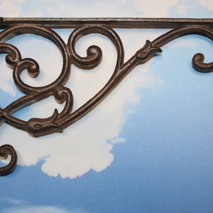 May include: Ornate, dark brown cast iron shelf bracket with a decorative scroll design. The bracket features a right-angle shape with a vertical support and a horizontal arm, perfect for supporting shelves. The design includes swirls and floral accents.