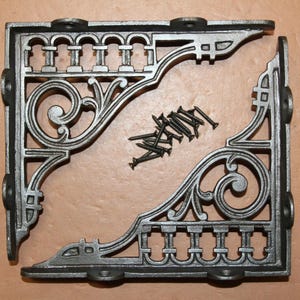 May include: Two ornate, cast iron corner brackets with a dark gray finish. Each bracket features a decorative scroll design and a series of small arches. A small pile of black screws is between the brackets. Hardware for home decor.