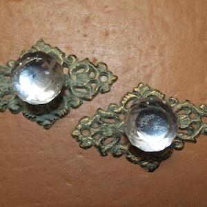 May include: Two drawer pulls featuring clear, faceted, round glass knobs mounted on ornate, diamond-shaped metal backplates with a distressed, dark bronze and gold-toned finish. Each backplate includes two small circular screw holes on the ends.