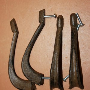 May include: A set of four dark brown, textured metal wall hooks with a curved, elongated design. Each hook features a mounting point at the top and bottom, and includes silver-colored metal screws for installation, showing a rugged finish.