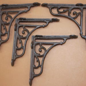 May include: Four dark brown cast iron shelf brackets. Each bracket has an ornate, scrolled design. The brackets are arranged in a group, with two facing one direction and two facing the opposite direction. The brackets are made of a heavy metal.