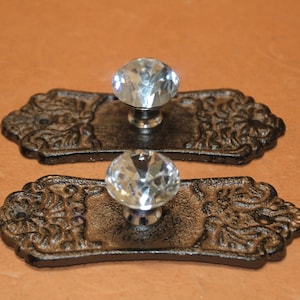 May include: Two drawer pulls featuring clear, multifaceted glass knobs attached to dark, textured, rectangular metal backplates. Each backplate displays an intricate, raised relief pattern on both ends and includes a small screw hole for mounting.