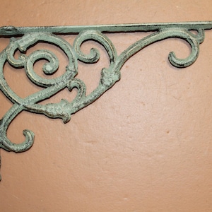 May include: An ornate, cast iron shelf bracket with a distressed, verdigris finish. The bracket features a right-angle design with decorative scrollwork and a curved support arm. Ideal for supporting shelves or decorative items.