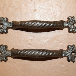 May include: Two dark brown metal drawer pulls featuring a twisted rope-style center grip and decorative fleur-de-lis ends. Each handle includes a circular screw hole at both extremities, showcasing a textured, weathered finish throughout the design.
