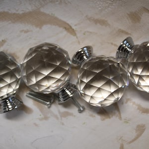 May include: Four spherical cabinet knobs made of clear, faceted glass that reflects light. Each knob features a polished silver-tone metal base and includes a threaded metal screw for installation, showcasing a geometric, multi-faceted surface design.