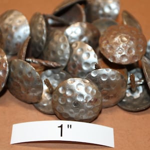 May include: A collection of antique-style, hammered metal upholstery tacks. The tacks have a domed head with a textured surface and a rusty, aged appearance. The label reads "1" inch.