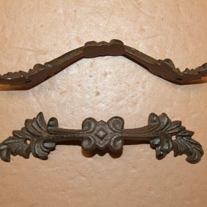May include: Two dark brown cast iron drawer pulls with an ornate, symmetrical leaf and scroll design. Each handle features a central diamond-shaped detail flanked by intricate foliage patterns and two mounting holes for attachment to furniture.