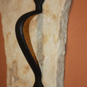 May include: A black metal handle with a curved design and pointed, spade-shaped ends featuring screw holes. The handle is mounted vertically onto a piece of rough, light-colored, textured natural stone with irregular edges and a flat surface.