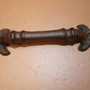 May include: A dark brown, textured cast iron drawer pull handle featuring a central cylindrical bar with two raised bands and decorative, pointed, fleur-de-lis style mounting plates at each end, each containing a single circular screw hole.