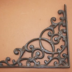 May include: A decorative, dark brown cast iron shelf bracket with an ornate, scrolled design. The corner bracket features a right angle and is suitable for supporting shelves or other decorative elements. The bracket is approximately 8 inches long and 8 inches wide.