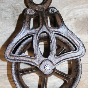 May include: A rustic, cast iron pulley with a wheel and a loop for hanging. The pulley is a dark brown color with a textured, aged appearance. The wheel has spokes and a central bolt. The design is symmetrical and industrial.