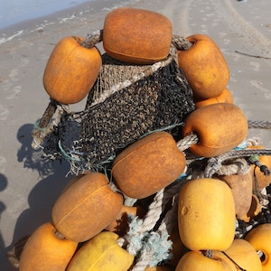 May include: A collection of orange and yellow fishing buoys with rope and netting. The buoys are various sizes and shapes, some with the word "SAWA" printed on them. The background is a sandy beach.
