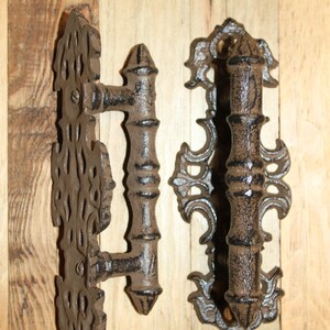 May include: Two dark brown, textured metal door handles featuring ornate, scrollwork backplates with intricate cutouts. Each handle has a central cylindrical grip with pointed ends and is secured to the decorative backplate with visible screws.