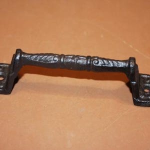 May include: A black cast iron door handle featuring a textured, cylindrical grip with decorative ridges and two flat, rectangular mounting plates at each end, each containing a single circular screw hole for secure installation on a surface.