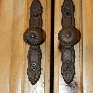 May include: Two dark brown, cast iron cabinet knobs with a round, fluted center and an elongated, decorative backplate featuring leaf-like detailing. Each knob is secured to a light-colored, natural wood cabinet door with two visible screws.