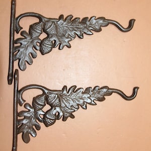 May include: Two cast iron plant hangers. Each hanger features an oak leaf and acorn design, with a curved hook for hanging. The hangers are dark brown and are mounted on a light brown surface.
