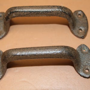 May include: Two dark gray, textured metal handles with a curved arch shape. Each handle features two flat, oval-shaped mounting plates at the ends, with two pre-drilled screw holes on each plate for secure attachment to a surface or door.