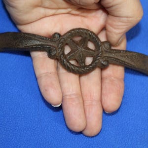May include: A dark brown cast iron handle featuring a central circular motif with a five-pointed star surrounded by a rope-like texture. The handle has two flat ends with circular holes for mounting, and the surface has a textured, rustic finish.