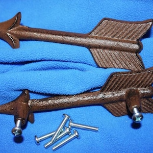 May include: Two brown cast iron arrow-shaped drawer pulls with a textured finish. Each pull features a pointed arrow head and a fletched tail end. The set includes several small silver-colored metal mounting screws for installation purposes.