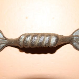May include: A dark gray cast iron drawer pull featuring a central twisted rope design flanked by two leaf-shaped ends. Each leaf end contains a circular hole for mounting hardware, providing a functional and textured grip for furniture cabinets.