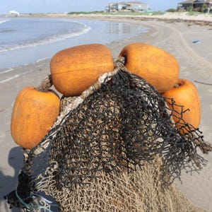 May include: Close-up of a fishing net with four orange buoys. The net is black and beige, with the buoys attached at the top. The background shows a sandy beach and the ocean.