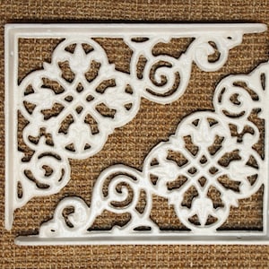 May include: Two white, ornate, cast iron corner brackets. Each bracket features a decorative floral and scroll design. The brackets are shown against a brown, textured background.