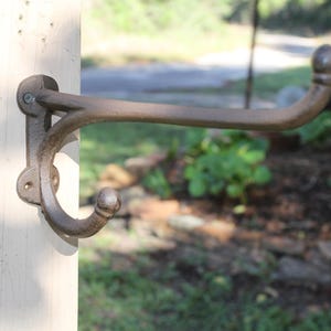 May include: A brown, cast iron wall hook with a double-pronged design, featuring a long upper arm and a shorter lower arm, each ending in a rounded knob. The hook is mounted to a vertical wooden post with two visible screws through its base plate.
