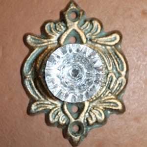 May include: A clear, faceted glass knob centered on an ornate, antiqued brass-toned metal backplate. The backplate features a symmetrical, raised leaf-like pattern with three circular screw holes positioned at the top, bottom, and center.