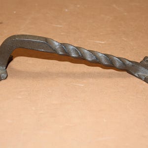 May include: A dark gray metal door handle featuring a twisted rope-like design in the center. The handle has a curved shape and includes two rectangular mounting plates at each end, each with visible screw holes for attachment to a surface.