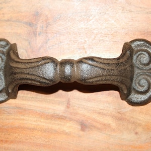 May include: A dark brown cast iron door handle featuring a textured surface and symmetrical scrollwork patterns on both ends. The handle has a central grip section and two circular mounting holes, one on each side, for attachment to a door or cabinet.