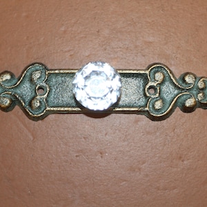 May include: A horizontal drawer pull featuring a weathered bronze-toned metal base with ornate scrollwork and two screw holes. A single, clear, faceted glass knob is centered on the metal plate, reflecting light with a bright, multi-sided appearance.