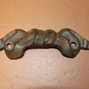 May include: A dark brown, cast metal drawer pull featuring a twisted, rope-like center design with two circular mounting holes and decorative, flared ends. The hardware has a textured, matte finish and a horizontal orientation.