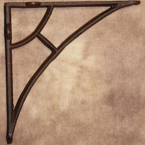 May include: A dark brown, cast iron shelf bracket with an ornate, openwork design. The bracket features a right-angle shape with a decorative curved support and a cross-shaped element. The metal has a textured surface and a rustic appearance.