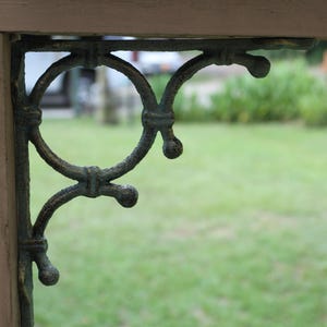 May include: Close-up of a decorative, antique-style metal bracket. The bracket is dark green with an ornate, swirling design and is attached to a wooden post. The background is blurred, showing green grass and foliage.