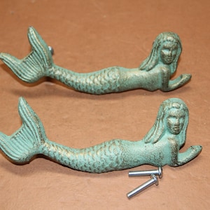 May include: Two horizontal mermaid-shaped drawer pulls with a weathered verdigris green and gold finish. Each handle features a detailed mermaid figure with a long tail and flowing hair. Two small silver-colored metal screws are included for installation.