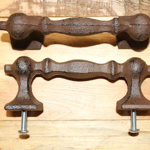 May include: Two dark brown cast iron drawer pulls with a textured finish. Each handle features a central bar with rounded ends and flared mounting feet. The bottom handle includes two silver-colored metal screws attached to the mounting points.