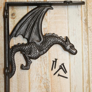 May include: A dark gray cast iron shelf bracket featuring a detailed dragon design with wings and scales, including four matching black screws for installation. The bracket has a vertical and horizontal arm with pre-drilled holes for mounting.