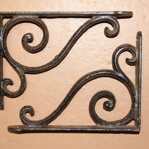 May include: Two ornate, black metal shelf brackets with a scrolled design. Each bracket has a flat base and a right-angle support with decorative swirls. The brackets are likely for home decor or shelving.