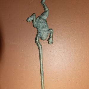 May include: A decorative metal hook in the shape of a frog. The frog is positioned as if climbing down the hook. The hook is a muted green color, with a curved top and bottom.