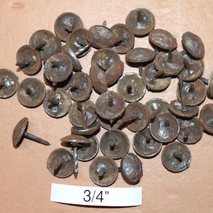 May include: A collection of antique, rusty-brown upholstery tacks with a 3/4" diameter. The tacks have a rounded head and a central pin, scattered on a light brown surface. The heads show signs of age and wear.
