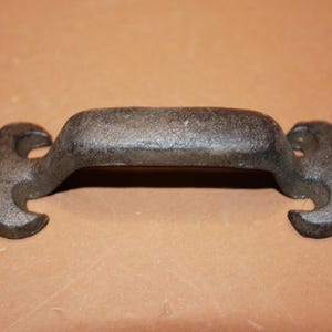 May include: A dark gray, cast iron drawer pull featuring a central horizontal handle and two decorative, pointed mounting plates on either end. Each mounting plate includes a single circular screw hole for attachment to a surface.