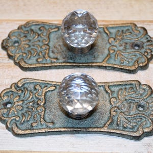 May include: Two drawer pulls featuring clear, multifaceted glass knobs centered on rectangular, ornate metal backplates. The backplates have a weathered, dark bronze and teal patina finish with intricate, raised scrollwork and two screw holes.