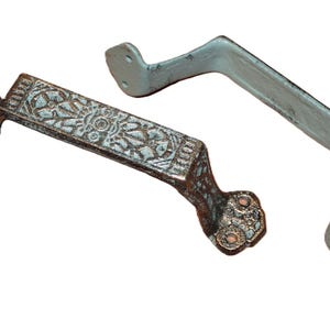 May include: Two metal drawer pulls with mounting plates at each end. One pull features a dark, weathered finish with an intricate embossed geometric floral pattern on the handle. The second pull has a smooth, solid light gray or off-white finish.
