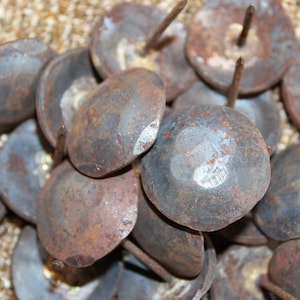 May include: A collection of antique, round upholstery tacks with a hammered, aged appearance. The tacks have a dark, metallic finish with visible rust and wear, and each has a central nail.