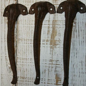 May include: Three dark brown cast iron door handles shaped like elephant heads with long trunks. Each handle features two circular screw holes located on the ears for mounting purposes, with a textured surface detailing the elephant's features.